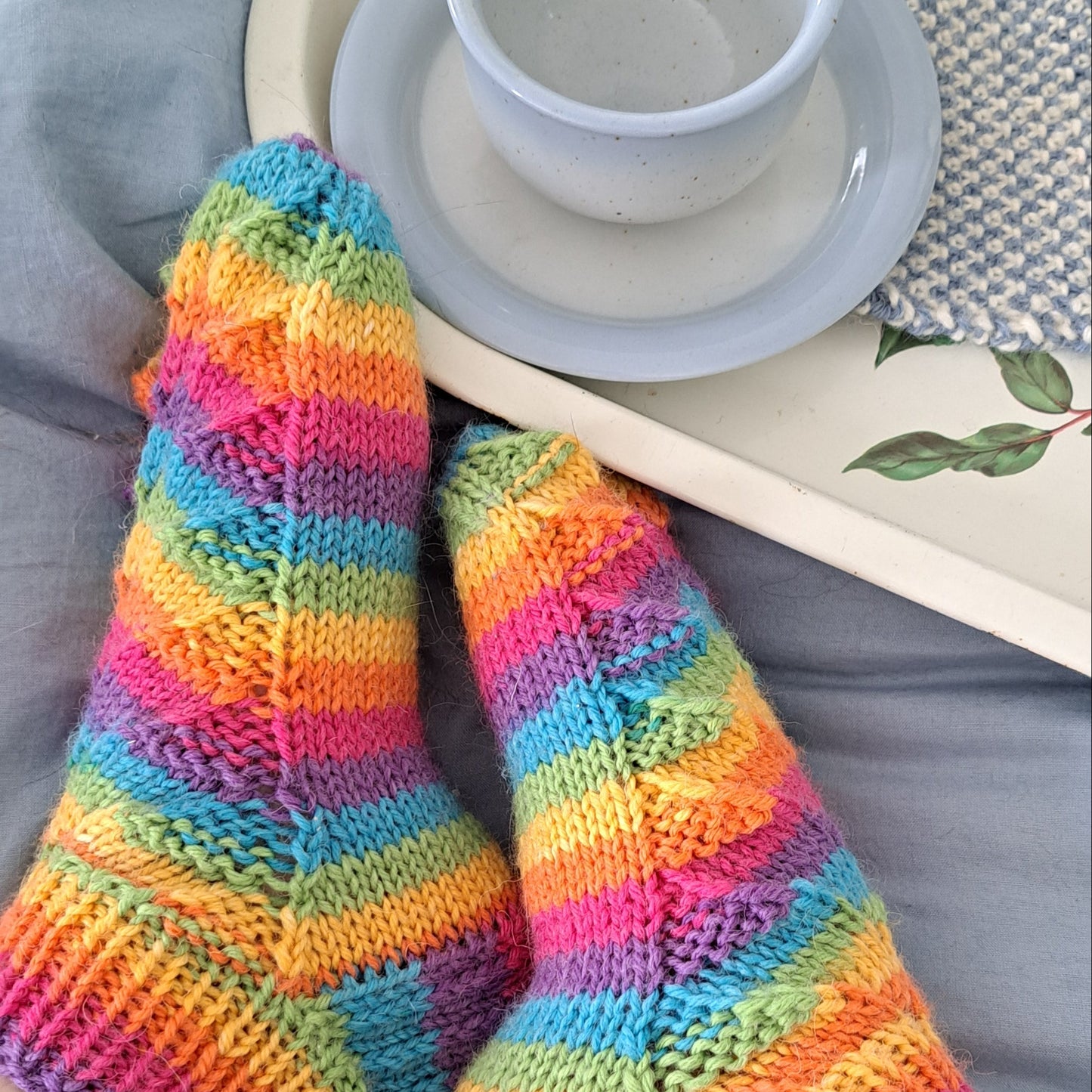 Paire de chaussettes arc en ciel tricotées main, près d'un plateau sur lequel est posé une tasse en céramique bleu ciel et un tapis de tasse bicolore bleu et blanc.